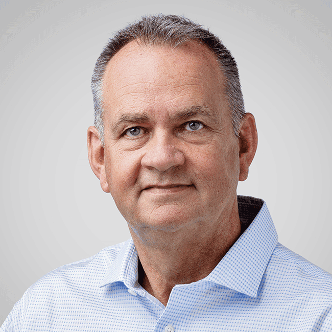 Portrait of a middle-aged man with short gray hair, wearing a light blue checkered shirt, against a neutral gray background.