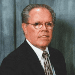 Portrait of a middle-aged man in a dark suit, white shirt, patterned tie, and glasses, looking at the camera with a neutral expression.