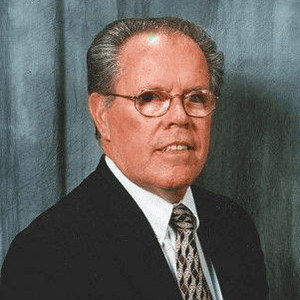 Portrait of a middle-aged man in a dark suit, white shirt, patterned tie, and glasses, looking at the camera with a neutral expression.