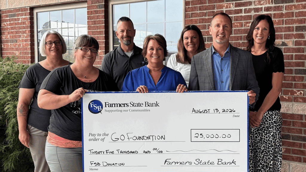 A group of seven adults in business casual clothing hold up a large ceremonial check while standing in front of a red brick building with windows