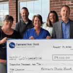 A group of seven adults in business casual clothing hold up a large ceremonial check while standing in front of a red brick building with windows