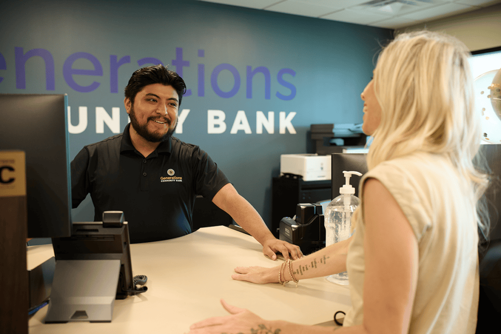 Bank teller smiles as a customer stands at the counter, sanitizer bottle on the desk and a teal wall with 'Generations' signage in the background.