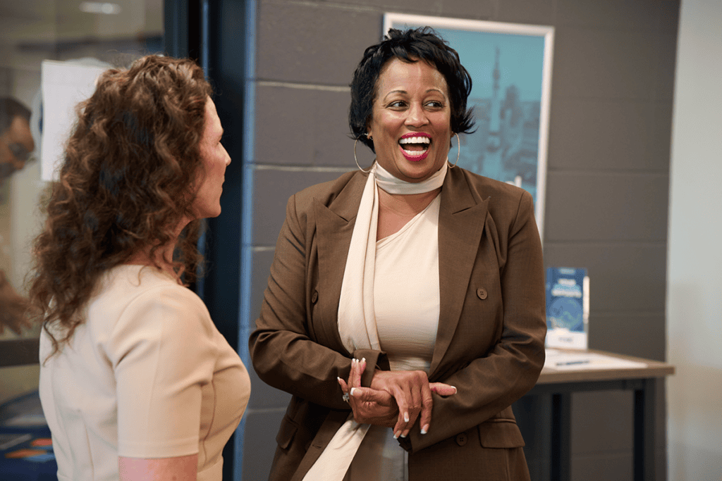 Two women in a modern office chatting and laughing, one wearing a brown blazer and scarf, the other in a light beige top; friendly conversation.
