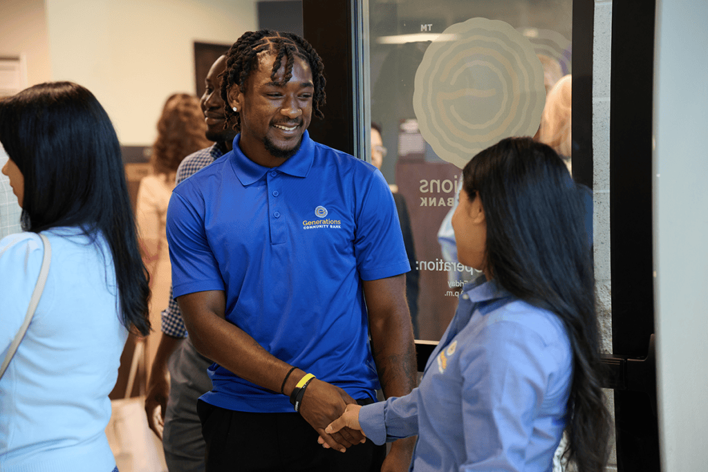Two people in blue polo shirts shake hands at a community bank event near a glass doorway, smiling at each other.