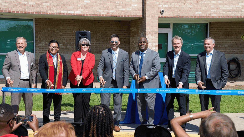 seven professionals in business dress clothing cut a blue ribbon in front of a brick building