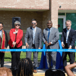 seven professionals in business dress clothing cut a blue ribbon in front of a brick building