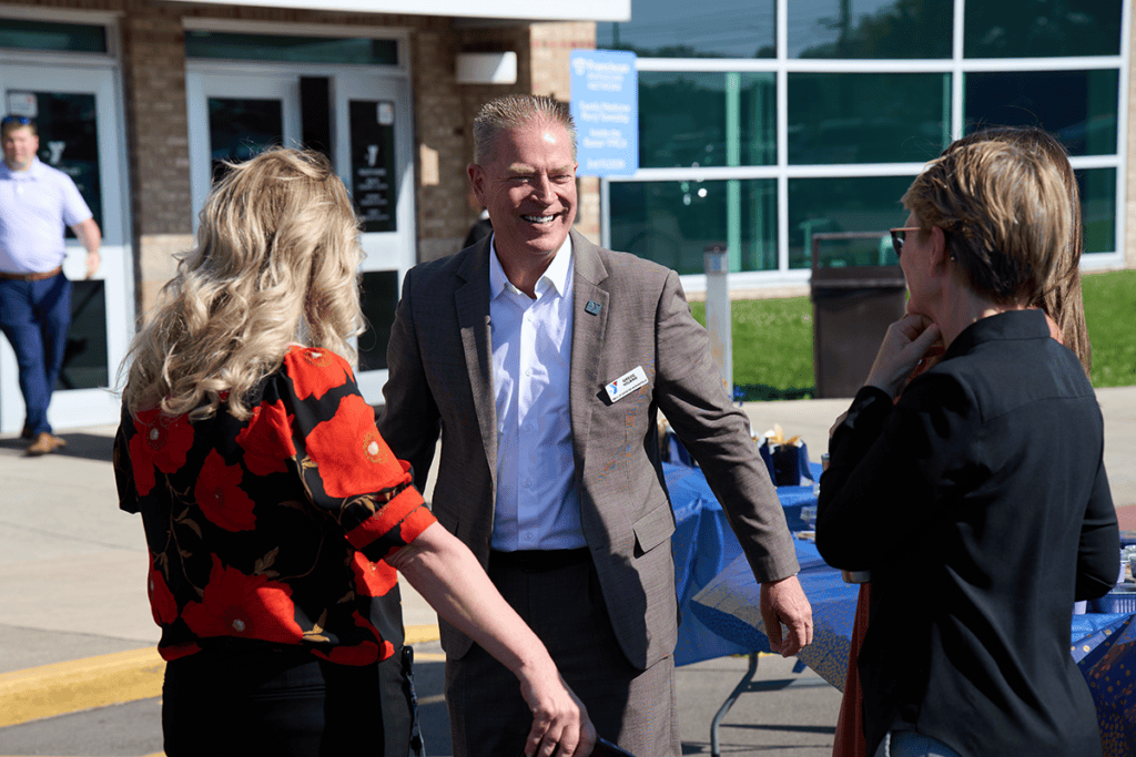 Businessman in gray suit chats with two women at an outdoor event near a table with blue tablecloths, smiling and engaging in conversation.