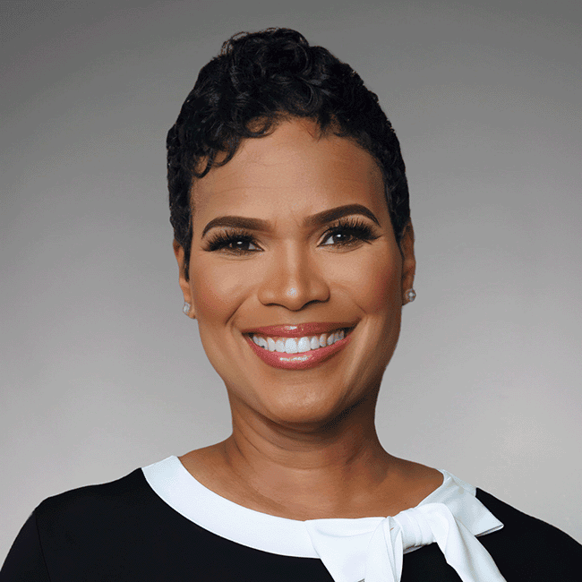 Smiling woman with short curly hair wearing a black top with a white bow, looking at the camera.
