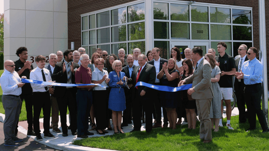 A photo showing a crowd of a couple dozen adults in professional clothing cutting a blue ribbon in front of a bank building.