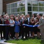 A photo showing a crowd of a couple dozen adults in professional clothing cutting a blue ribbon in front of a bank building.
