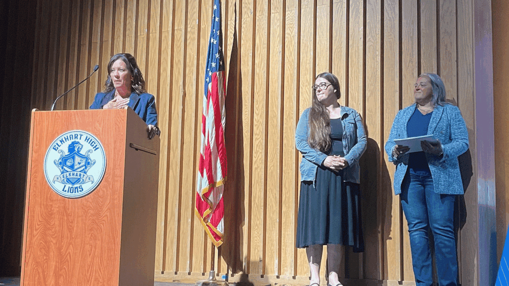 Woman at a wooden podium giving a speech on a stage; Elkheart High Lions crest on the podium and an American flag nearby.
