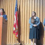 Woman at a wooden podium giving a speech on a stage; Elkheart High Lions crest on the podium and an American flag nearby.