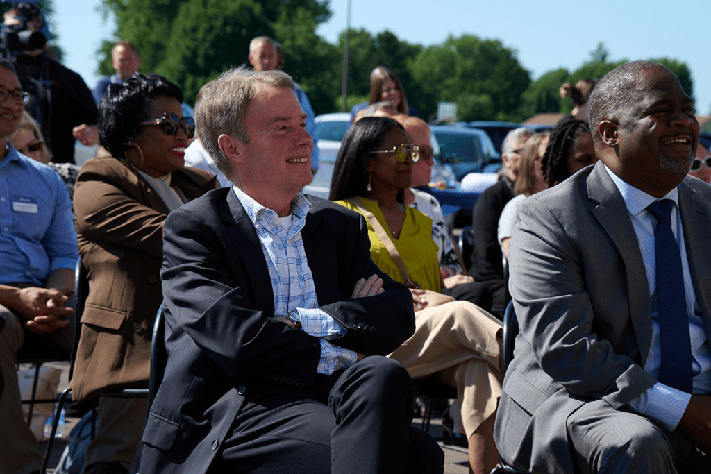a group of adults sit in folding chairs outside on grass under bright sunshine, looking at speakers at a podium