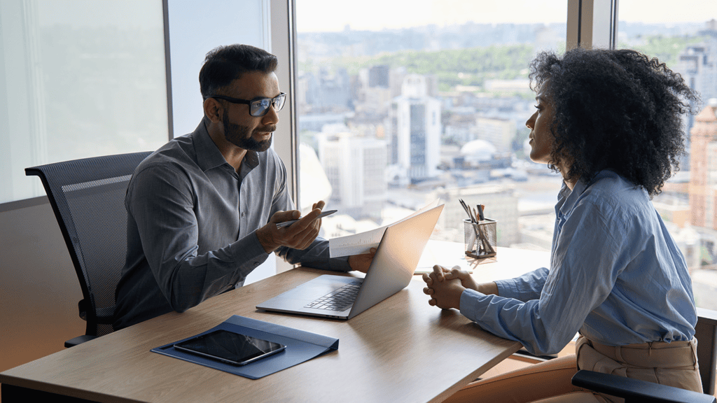 Two professionals in a glass-walled office discuss with a laptop open on the desk and a tablet nearby, city skyline in the background.