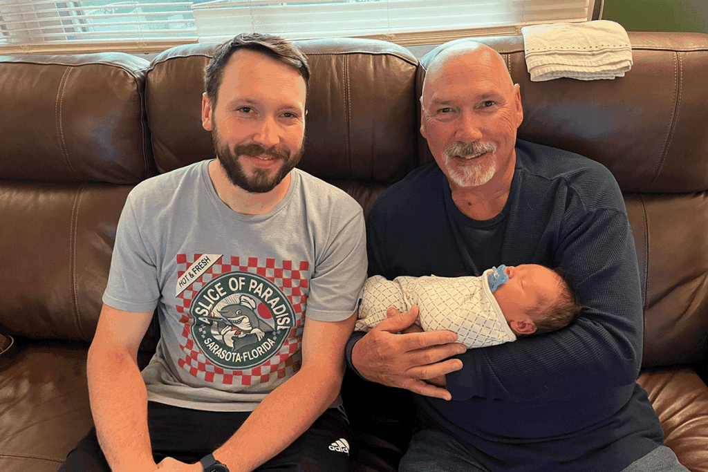 An older man holds his grandson while sitting on a couch next to his son, the baby's father
