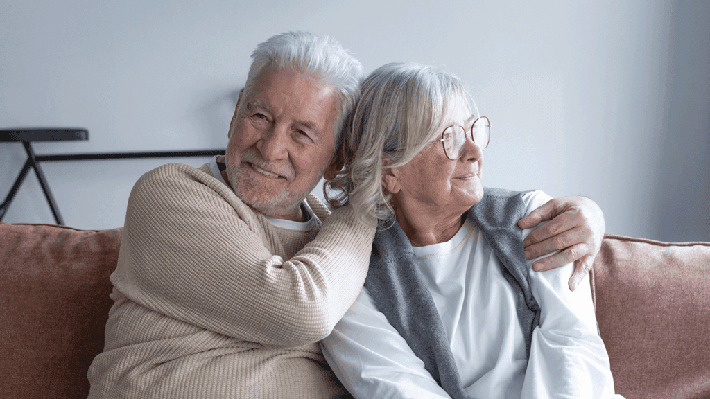Elderly couple sits close on a sofa, smiling and embracing with gentle affection