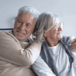 Elderly couple sits close on a sofa, smiling and embracing with gentle affection