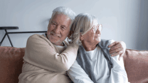 Elderly couple sits close on a sofa, smiling and embracing with gentle affection